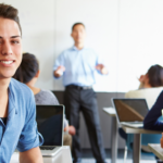 Young man smiling in classroom with teacher and laptops, reflecting A-Plus HVAC educational engagement and home solutions.