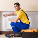 Young technician in yellow t-shirt and blue overalls performing HVAC maintenance on a radiator with an open toolbox.
