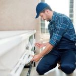 Male technician in blue cap and checkered shirt working on plumbing equipment by a bathtub in a sunny indoor setting.