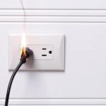 Electrical outlet with spark and black power cord on white wall, highlighting electrical safety concerns in home repairs.