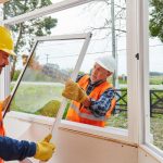 Two construction workers installing a window at a site, wearing safety helmets and high-visibility vests, near greenery.