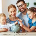 Family teaching kids about saving money with a piggy bank in a bright indoor setting.