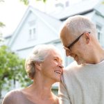 Older couple smiling in casual attire near a light house, representing companionship for aging homeowners.