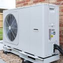 Air conditioning unit with fan and wheels, set against a brick wall and grass, by A-Plus Air Conditioning & Home Solutions.