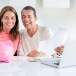 Smiling couple discussing energy-efficient upgrades at a table with a piggy bank and documents, near a laptop.